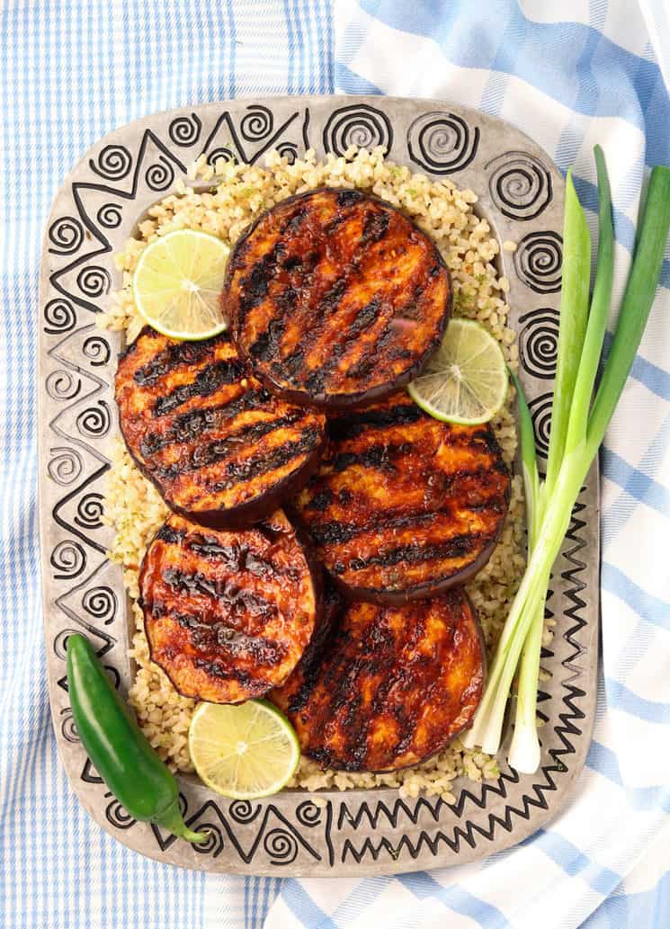 A platter of spicy grilled eggplant on coconut lime rice, with limes, jalepeno and green onion, on a blue plaid towel