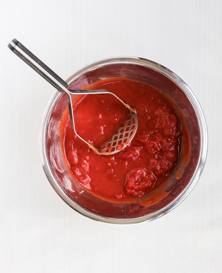 smashed whole tomatoes in a metal bowl with a potato masher.