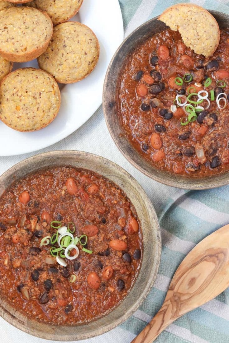 overhead shot of Best damn Vegan Chili in 2 wooden bowls and a wooden spoon with polenta muffins on a green striped towel.