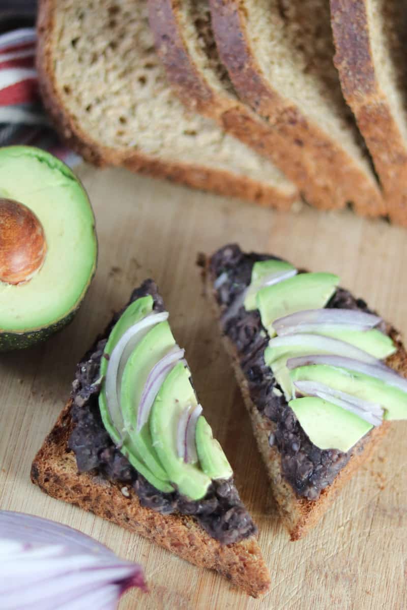 Smashed Black Beans Avocado Toast on a cutting board with red onion, bread, avocado half. 
