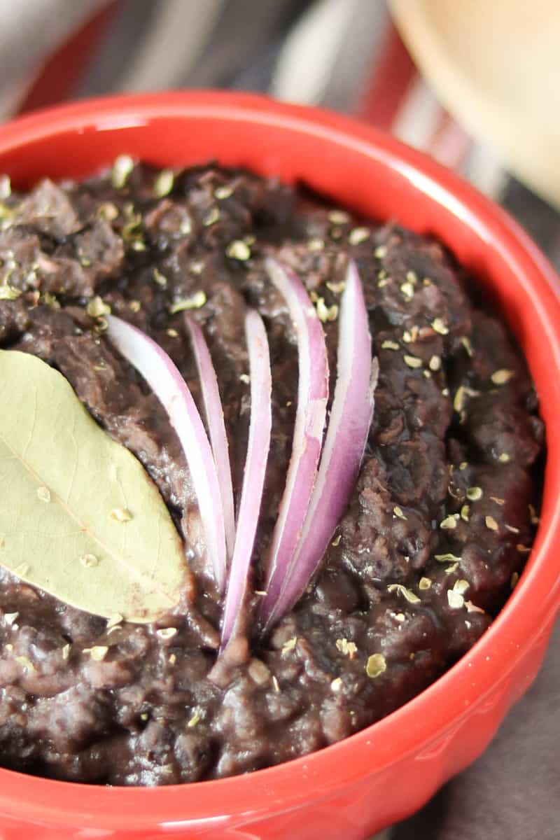 slow cooker black beans close up of black beans, a bay leaf and red onion in a red bowl on a striped towel with a wooden spoon