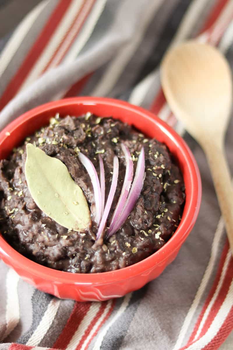easy slow cooker black beans black beans, a bay leaf and red onion in a red bowl on a striped towel with a wooden spoon