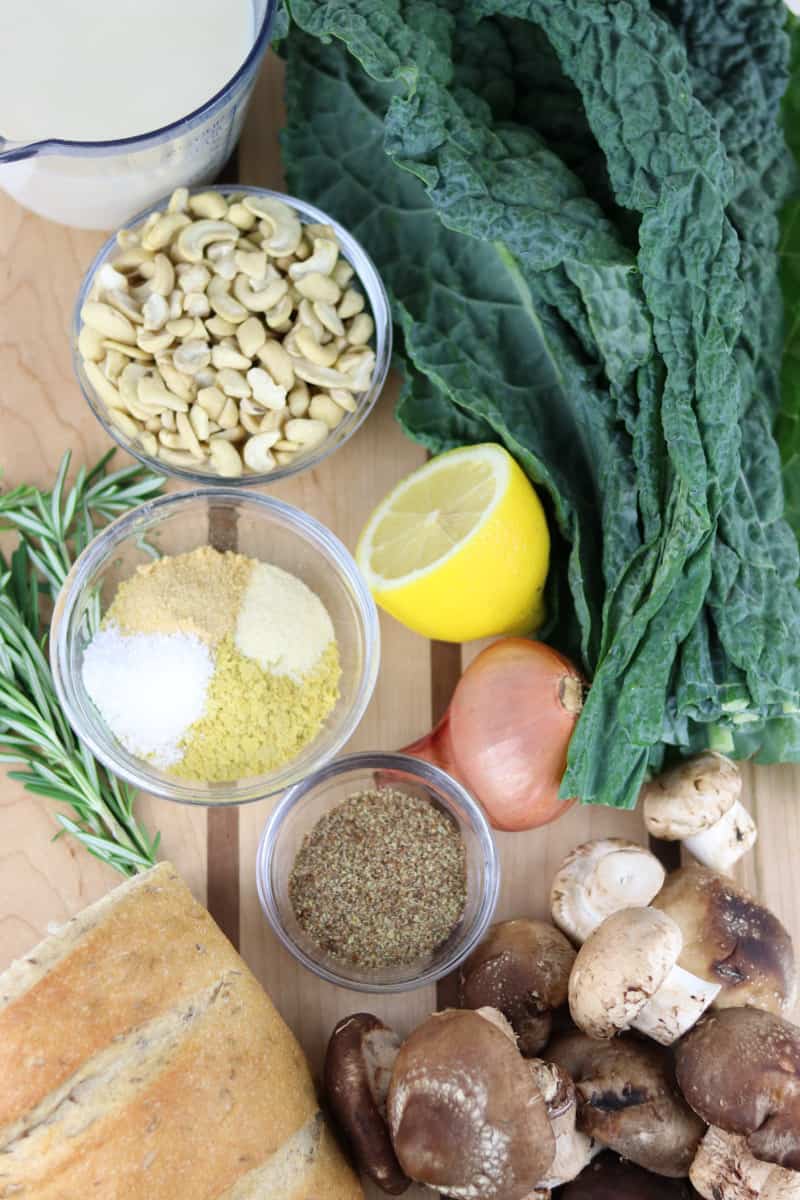 Kale Mushroom Bread Pudding Ingredients on a cutting board -- kale, cashews, plant milk, loaf of bread, lemon, shallot, mushrooms and seasonings.