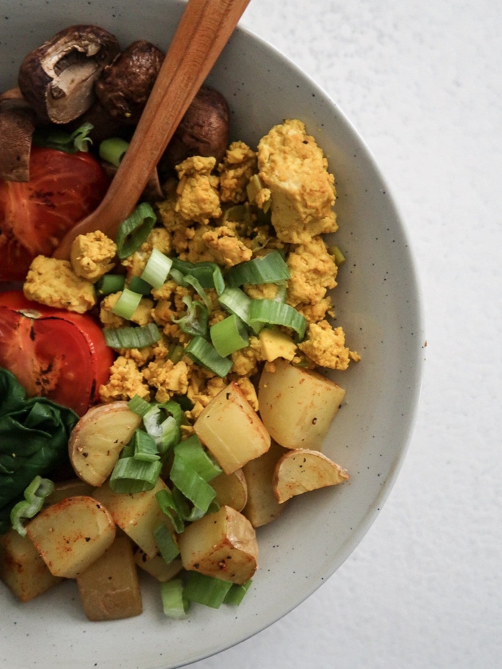 Tofu scramble in bowl with potatoes, green onions, mushrooms and tomatoes with spoon.