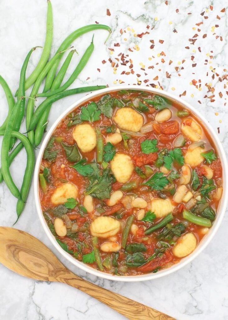 White bowl of tomato based gnocchi soup with green beans, white beans, herbs and crushed red pepper flakes on white marble.