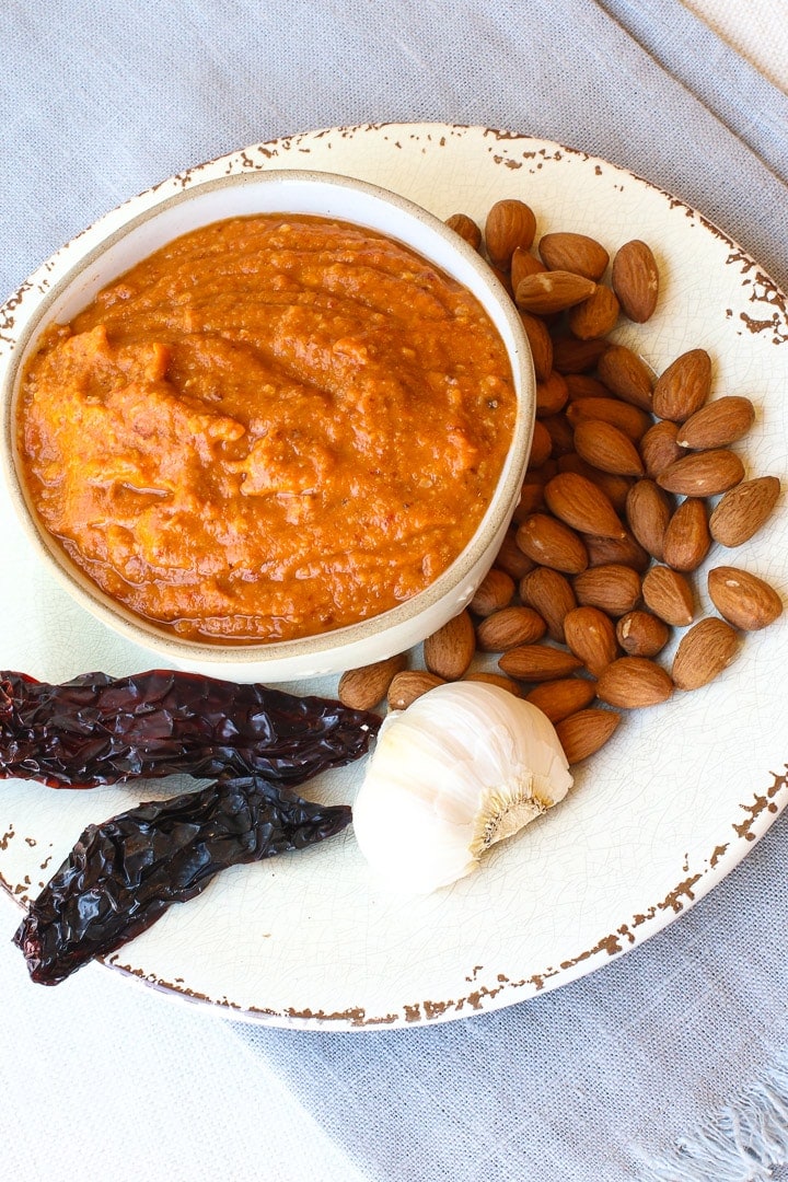 Romesco Sauce in a bowl on a white plate and blue towel with chilis, almonds and dried peppers