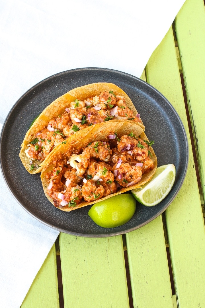 Cauliflower Tacos with Romesco Sauce on a dark gray plate with limes on a lime green table