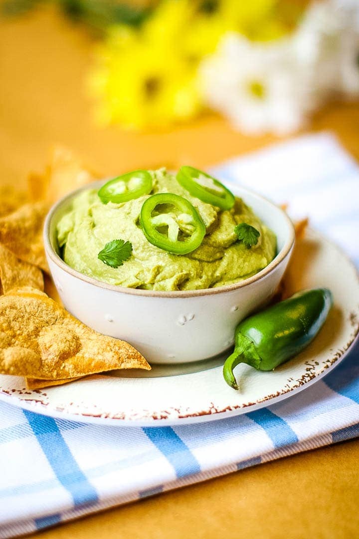 Spicy oil free hummus in a bowl with tortilla chips and jalapeño on a blur plaid towel with daisies