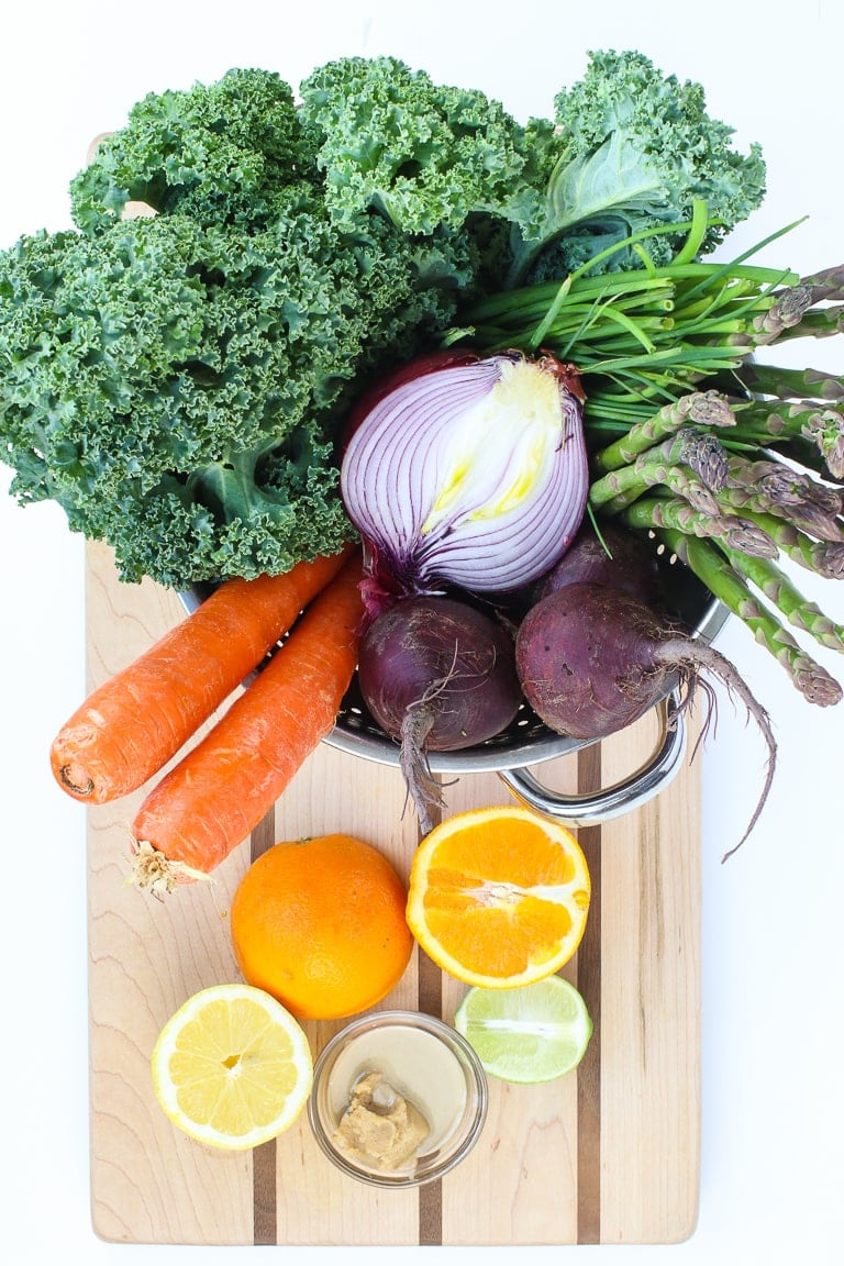 warm kale salad ingredients on a cutting board, kale, red onion, carrots, beets, lemon, lime, orange, miso, tahini