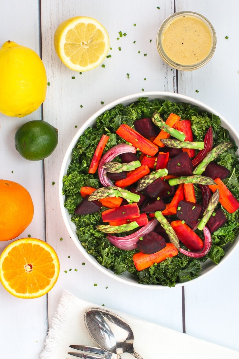 Warm kale salad with roasted carrots, beets and asparagus and citrus dressing on a white table with lemons, oranges and lime