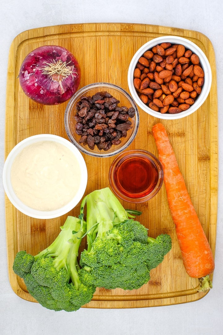 cutting board with broccoli, red onion, carrot, and dishes of mayo, maple syrup & rice vinegar, raisins and smoky almonds.