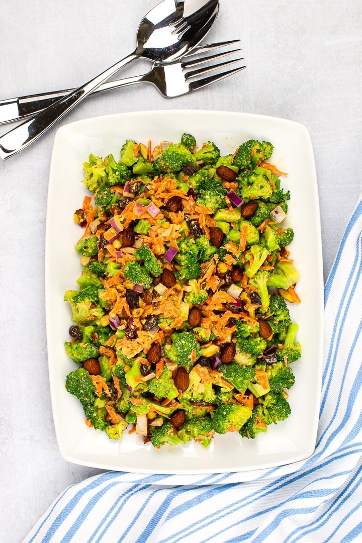 vegan broccoli salad with shredded carrots, diced red onion, raisins, smoky almonds and sweet & creamy dressing on a white platter with a blue striped napkin and fork. 