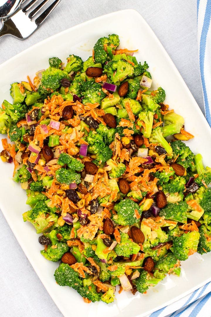 vegan broccoli salad with shredded carrots, diced red onion, raisins, smoky almonds and sweet & creamy dressing on a white platter with a blue striped napkin and fork. 