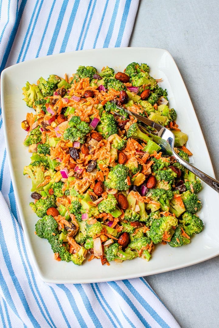 vegan broccoli salad with shredded carrots, diced red onion, raisins, smoky almonds and sweet & creamy dressing on a white platter with a serving fork in the salad on a blue striped napkin. 
