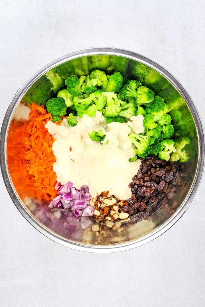 metal mixing bowl with blanched broccoli, shredded carrots, diced red onion, raisins, smoky almonds and sweet & creamy dressing.
