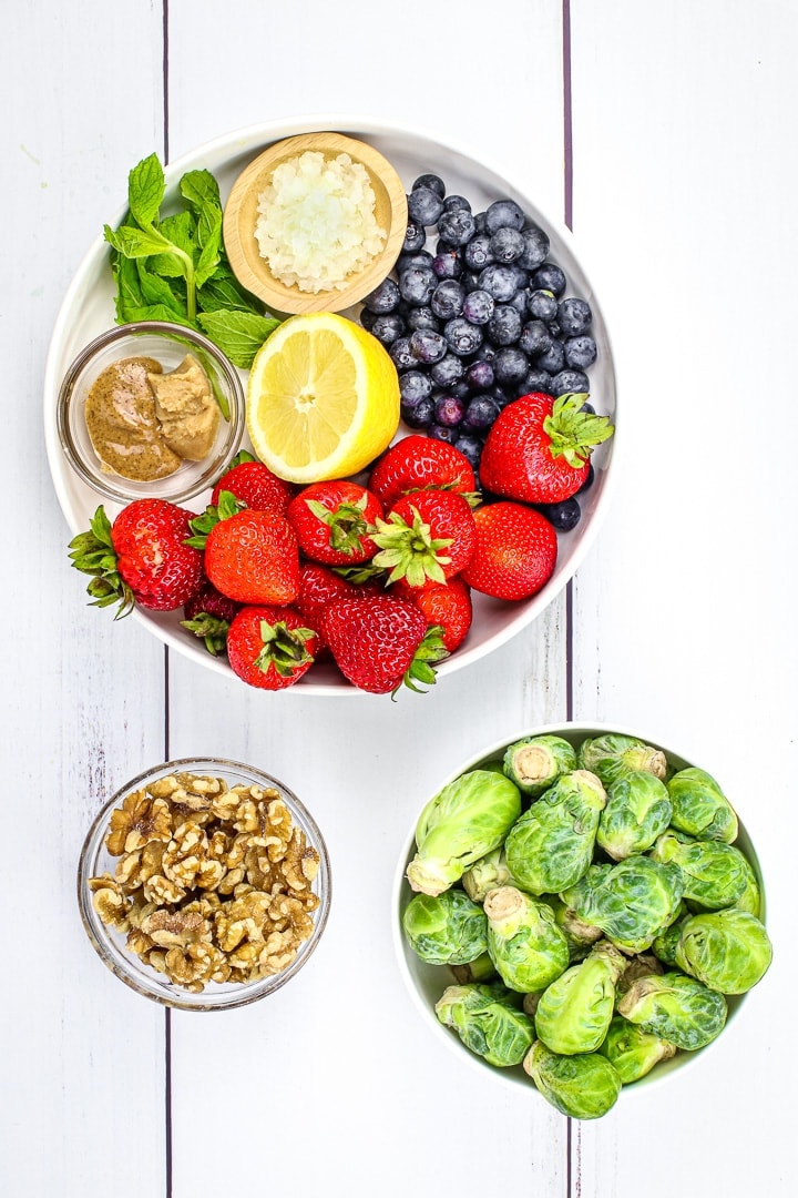 salad ingredients in bowls: strawberries, blueberries, lemon, miso paste, almond butter, mint leaves, diced shallot, brussels sprouts and walnuts