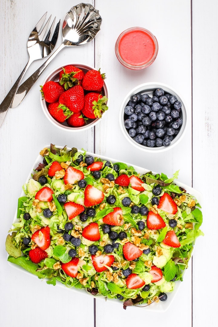 Shaved brussels sprouts, sliced strawberries, blue berries, mint leaves chopped walnuts on a bed of spring greens on a white platter with strawberry vinaigrette in a jar and bowls of berries.