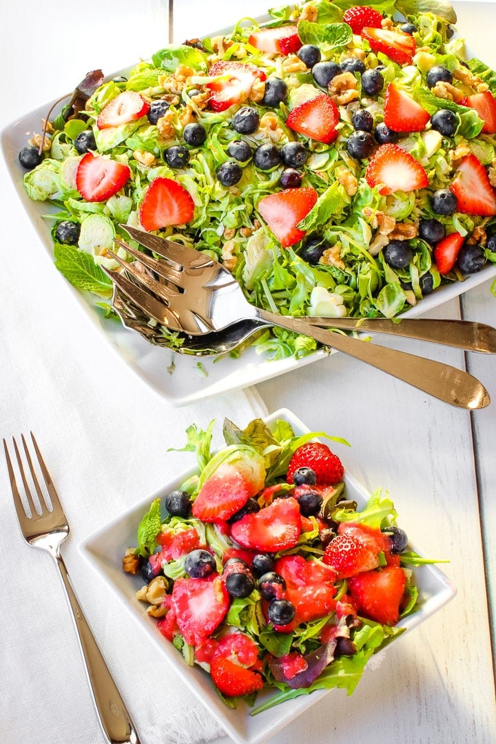 Shaved brussels sprouts, sliced strawberries, blue berries, mint leaves chopped walnuts on a bed of spring greens on a white platter with a small plate of salad and a fork