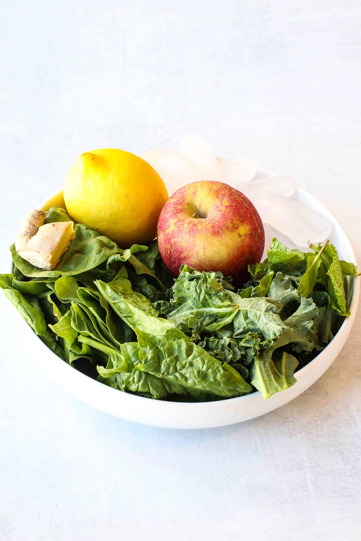 Green smoothie ingredients in a white bowl-apple, lemon, kale, spinach, ginger and ice cubes