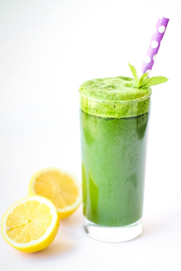 Green smoothie with ginger, in a tall class with cut lemons, mint garnish, and purple polka dot straw