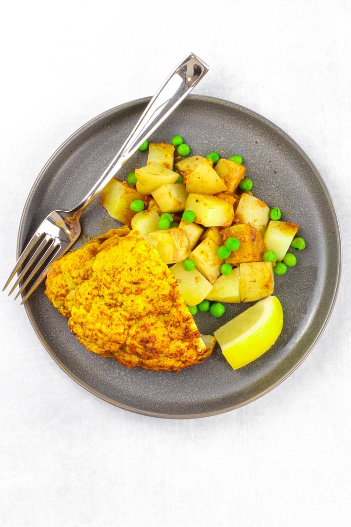 whole roasted cauliflower tandoori with potatoes and peas and lemon wedges on a dark gray plate with a fork 