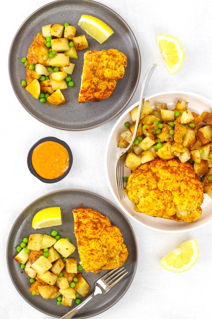 whole roasted cauliflower tandoori with potatoes, peas and lemon wedges on dark gray plates and in a white serving dish. A dish of sauce on the side.