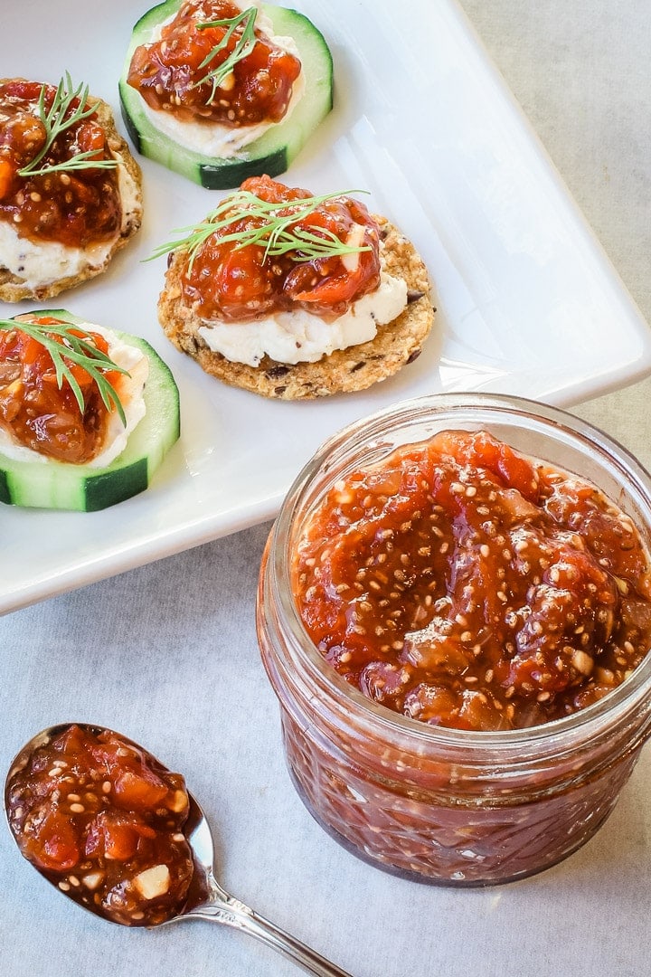 close up of tomato jam in a jar (with seeds) and on a small spoon, and white platters with tomato jam and nut cheese on cucumbers and crackers with dill garnish. 