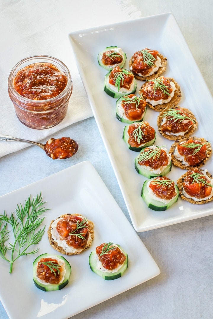 Tomato jam in a jar and on a small spoon, and white platters with tomato jam and nut cheese on cucumbers and crackers with dill garnish. 