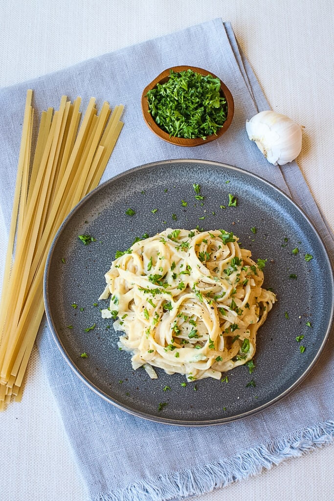 Blue Napkin with Gray plate with vegan fettuccine alfredo topped with herbs, with Bowl of herbs, dry pasta and garlic.