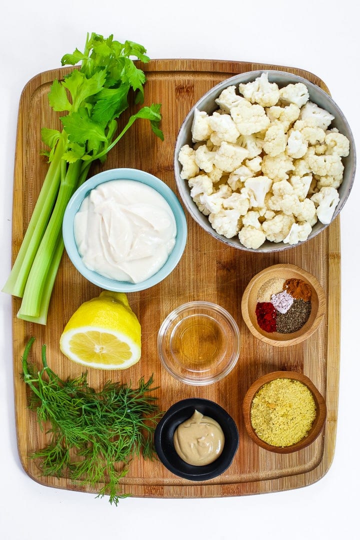 cutting board with celery, cauliflower florets, bowl of mayo, lemon half, dill sprigs, vinegar, dijon mustard, nutritional yeast and spices.