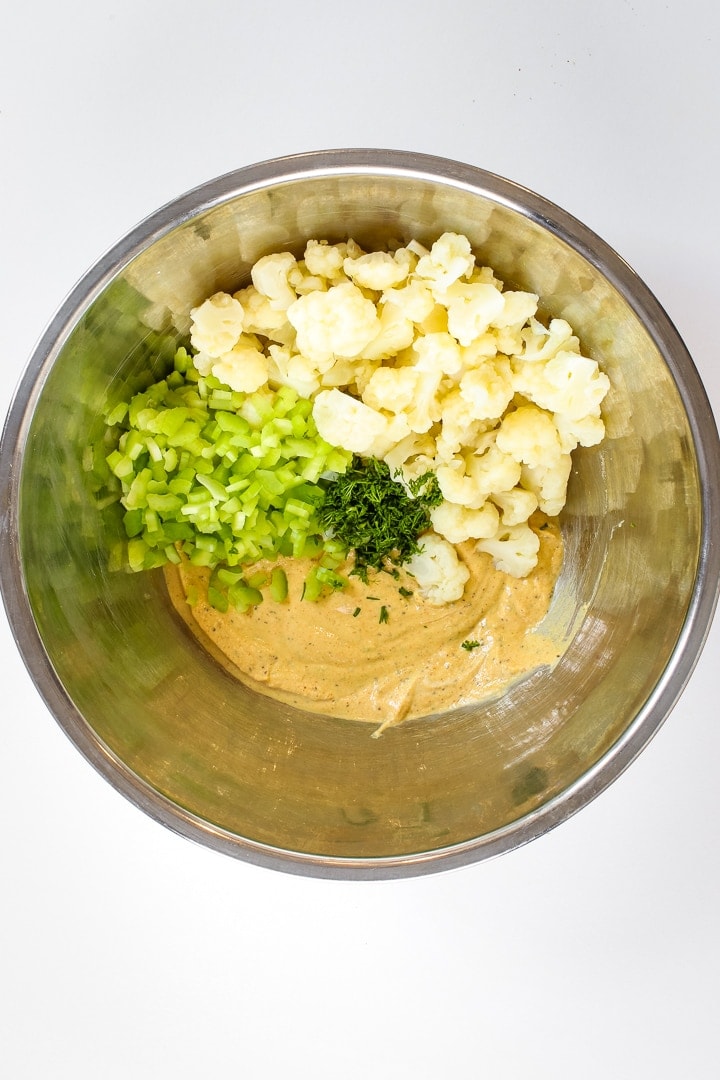 stainless steel mixing bowl of dark yellow dressing, diced celery, cauliflower florets and chopped dil