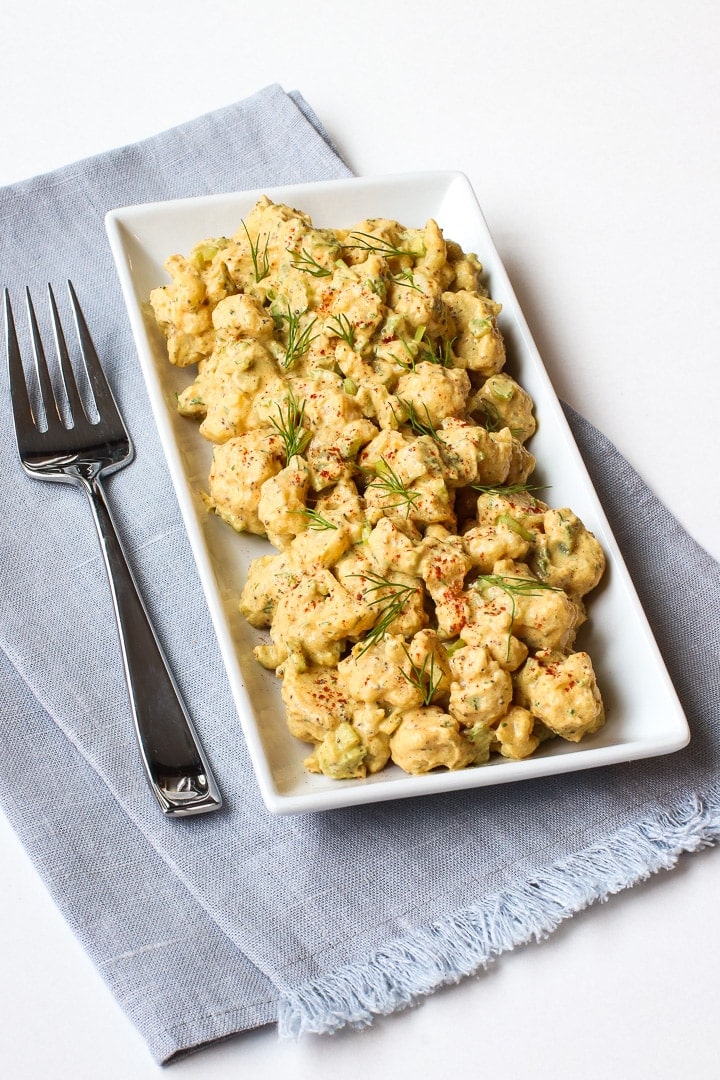 Cauliflower potato salad garnished with smoked paprika and dill sprigs, on a white rectangular dish on a blue towel with fork.