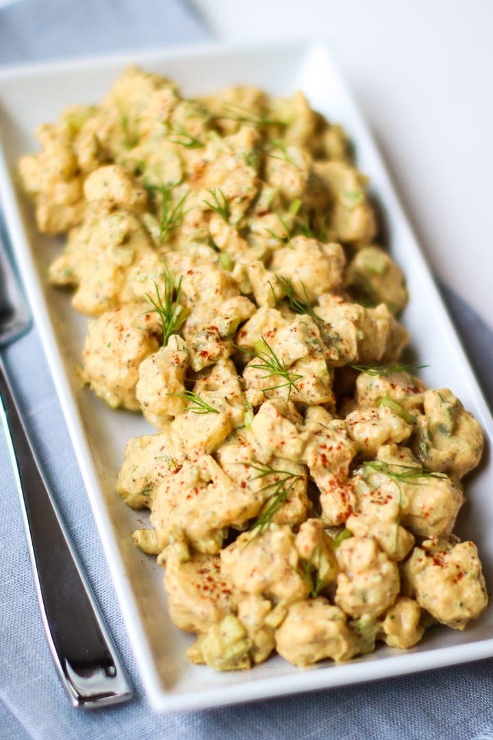 close up of Cauliflower potato salad garnished with smoked paprika and dill sprigs, on a white rectangular dish on a blue towel with fork.