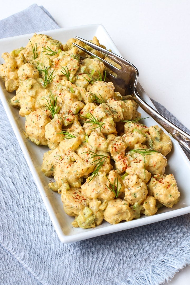 Cauliflower potato salad garnished with smoked paprika and dill sprigs, on a white rectangular dish on a blue towel with fork.