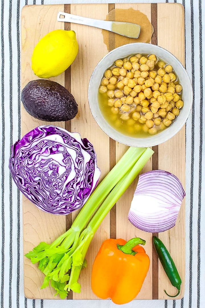 Bowl of chickpeas, lemon, tahini, celery, red onion half, purple cabbage half, avocado, orange bell pepper and serrano pepper on a striped wood cutting board on a black and white striped linen towel.