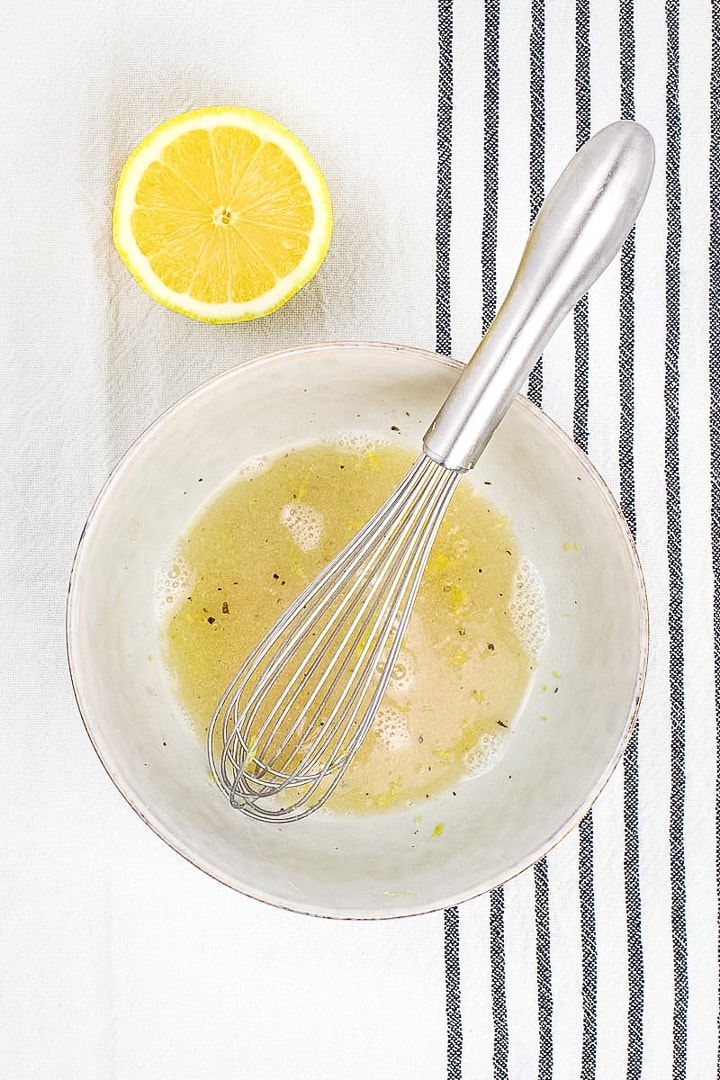 lemon aquafaba dressing in a light gray bowl with whisk and lemon half on black and white striped towel.