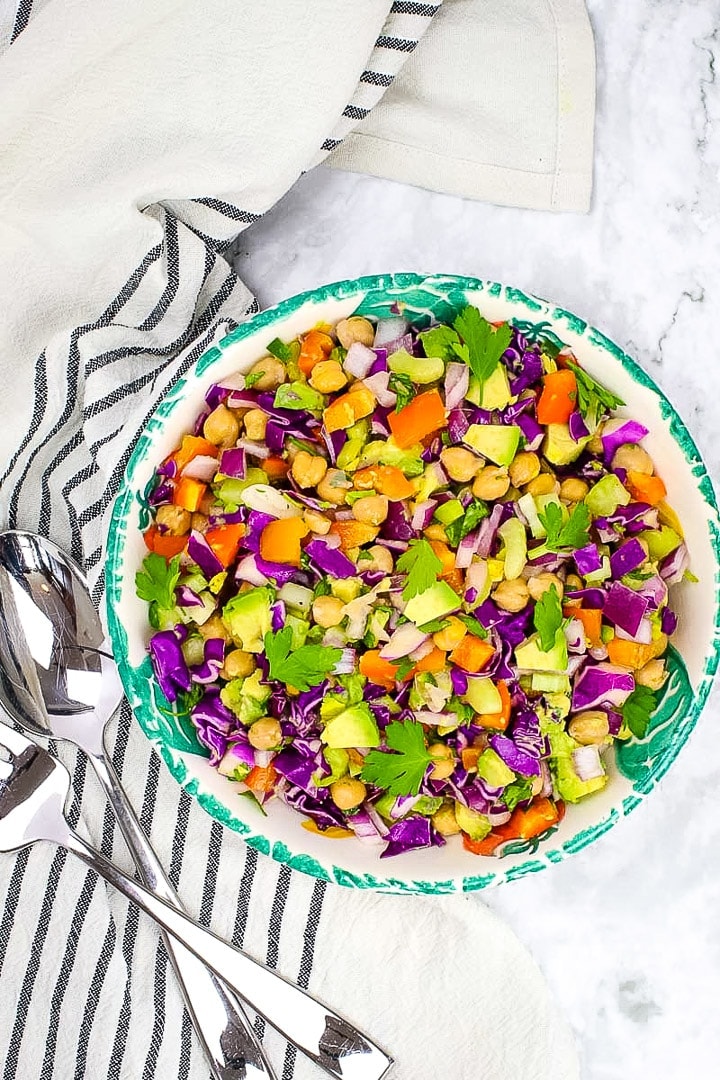 Simple chickpea salad in a wide bowl with green leaf trim, on marble with crossed fork and spoon and linens towel with black stripes.