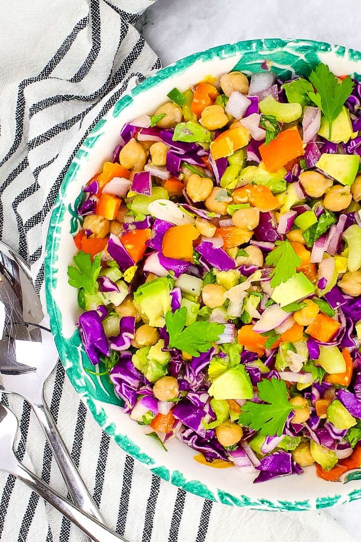 Simple chickpea salad in a wide bowl with green leaf trim, on marble with crossed fork and spoon and linens towel with black stripes.