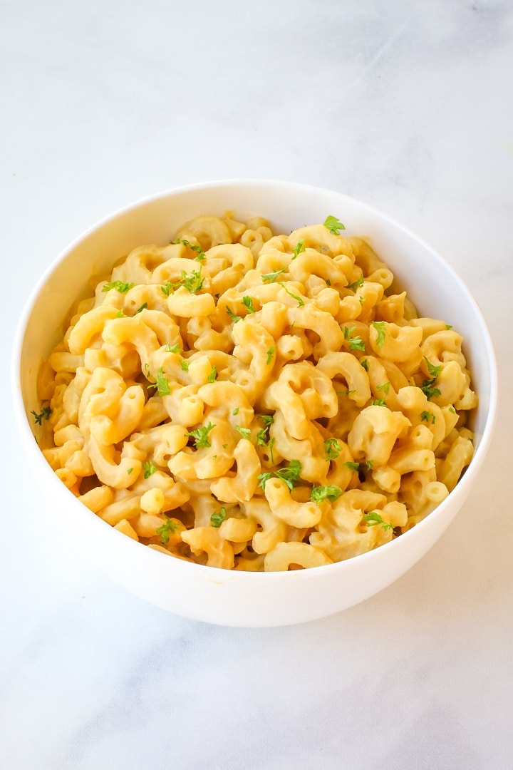 Elbow macaroni and creamy chickpea cheese sauce in a white bowl on white marble, topped with herbs.