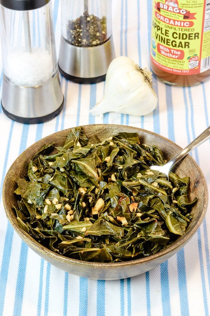 collard greens with garlic and smoked paprika in a wooden bowl on a pale blue striped towel with salt and pepper, garlic and apple cider vinegar.