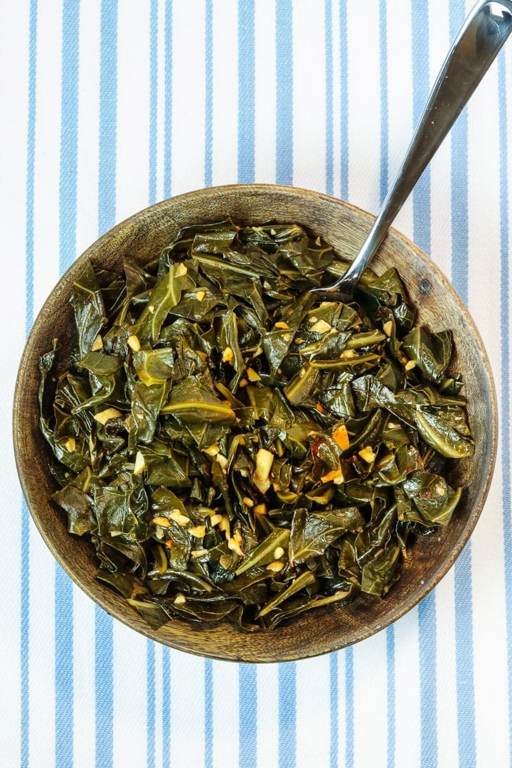 collard greens with garlic and smoked paprika in a wooden bowl with a fork on a pale blue striped towel.