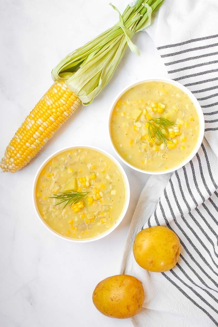White bowl of Lemony sweet corn chowder garnished with dill sprig on a marble background with a white towel with black stripes, potatoes, and an ear of corn. 