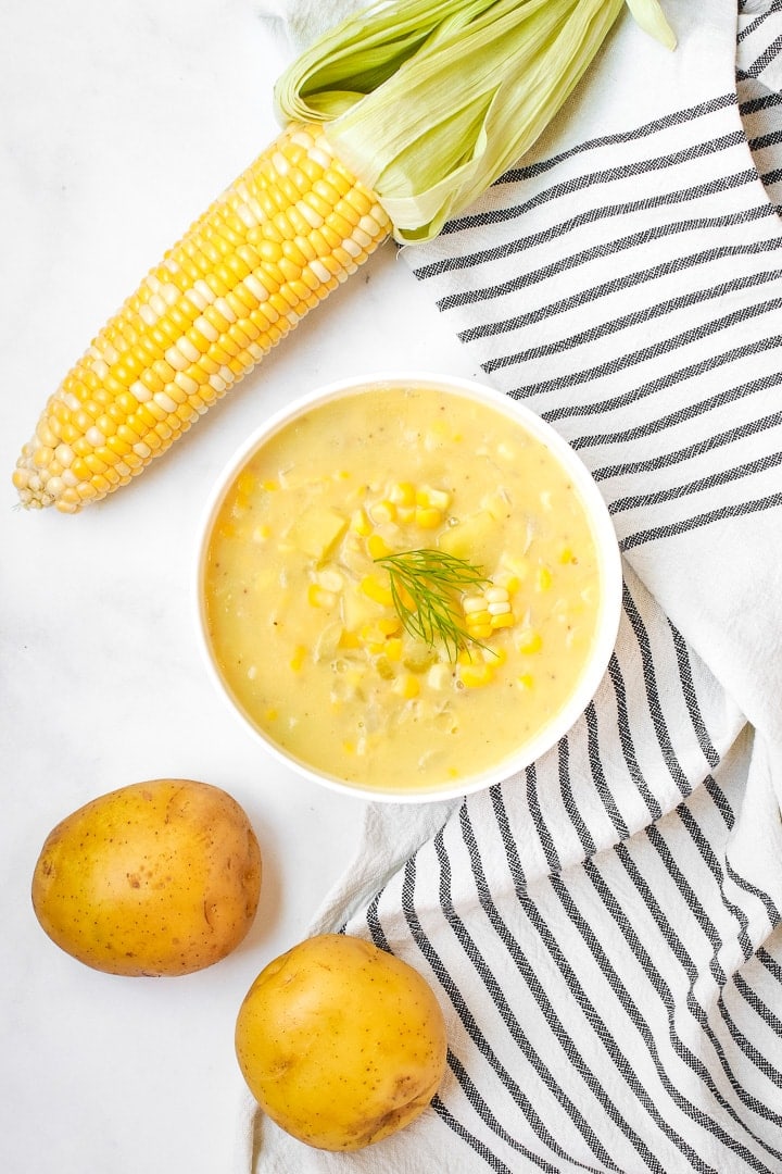 White bowl of Lemony sweet corn chowder garnished with dill sprig on a marble background with a white towel with black stripes, potatoes, and an ear of corn. 