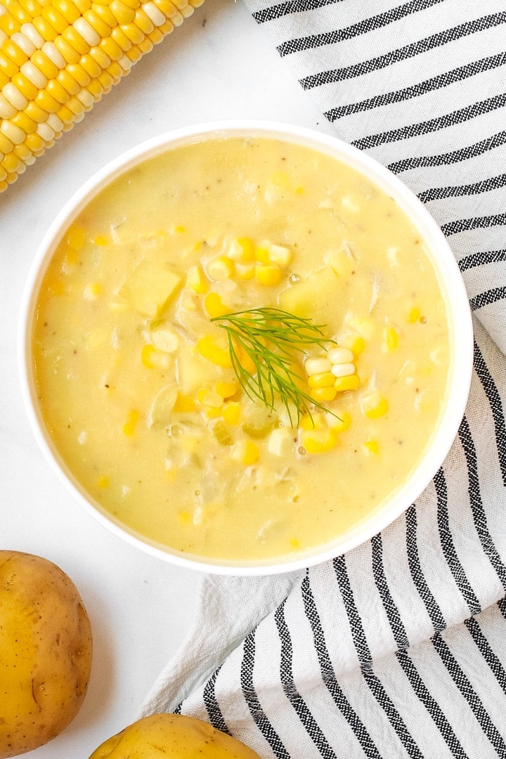Close up of white bowl of Lemony sweet corn chowder garnished with dill sprig on a marble background with a white towel with black stripes, potatoes, and an ear of corn. 