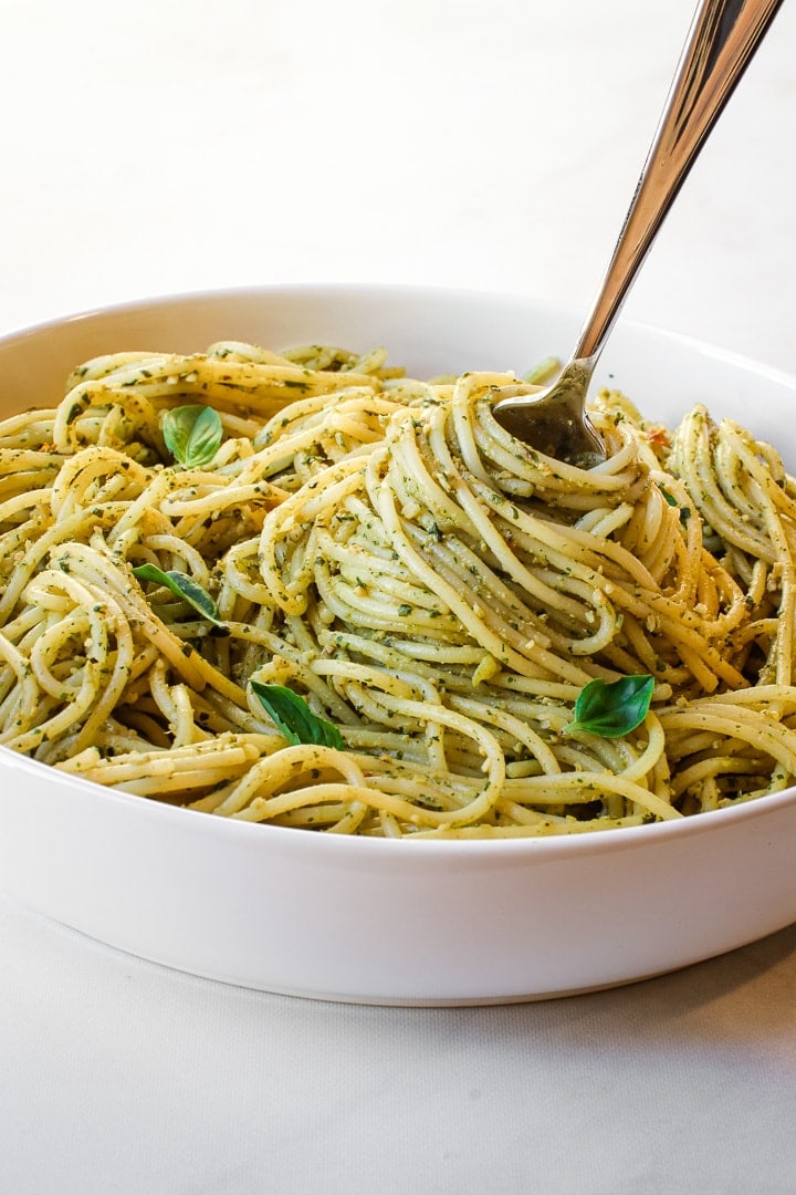 White bowl of spaghetti with pesto sauce and basil leaves with a pasta-filled twirling fork on marble.