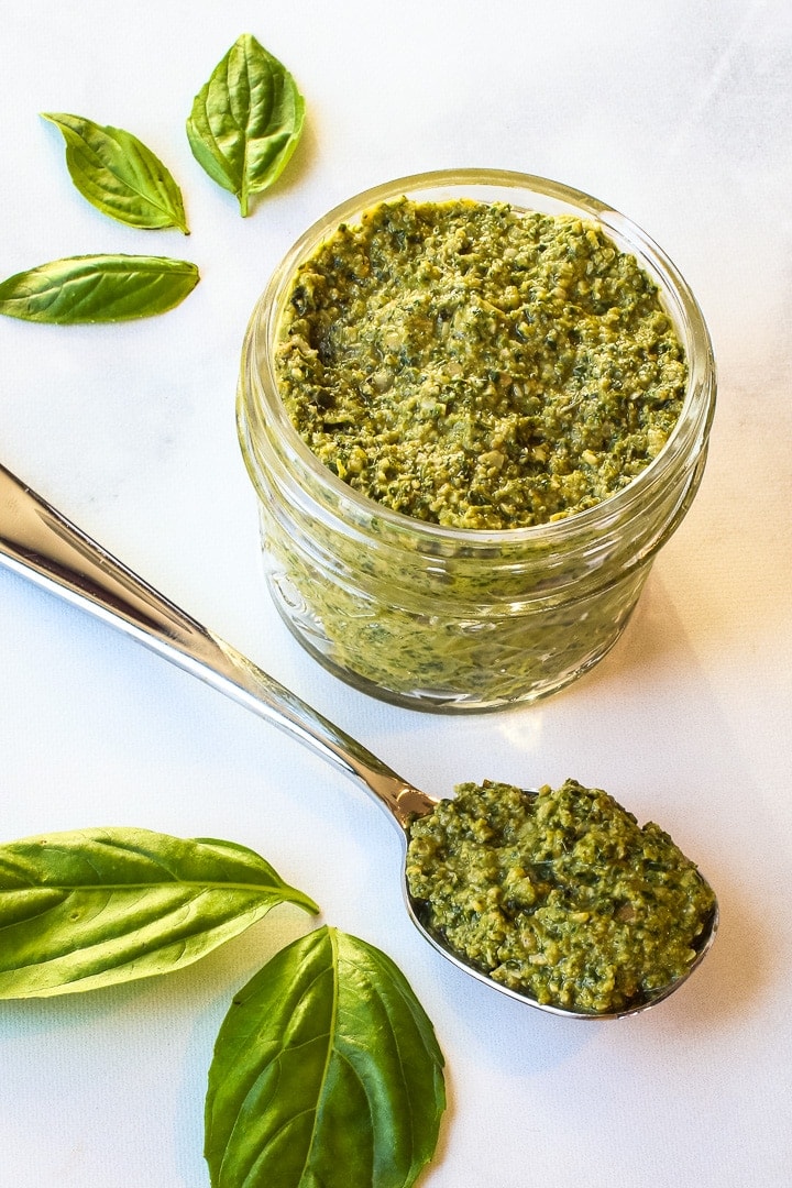 Jar of pesto and spoon of pesto with basil leaves on marble.
