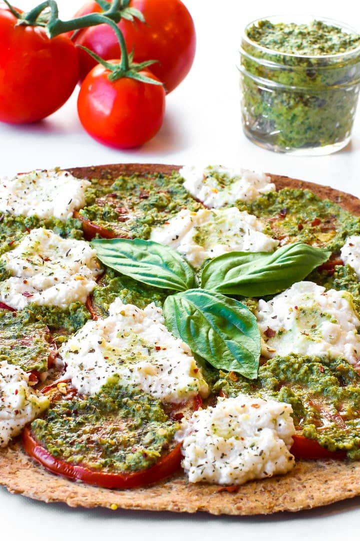 Thin crust pesto pizza with sliced tomatoes, fresh almond ricotta, seasonings and 3 basil leaves in center. Vine tomatoes and jar of pesto in background.