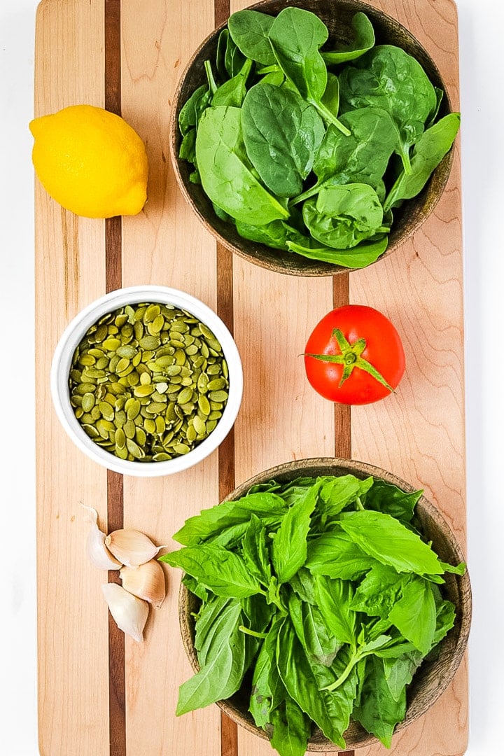 Striped cutting board with wooden bowls of spinach and basil, a lemon, tomato, garlic, and a white bowl of pepitas.