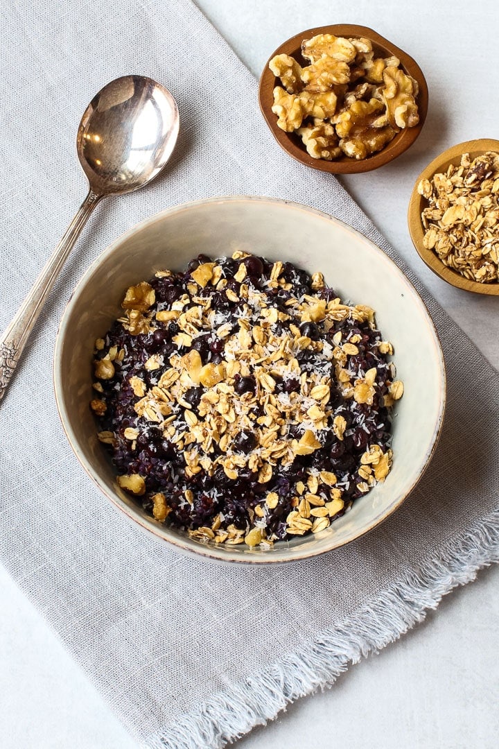 Bowl of quinoa porridge with blueberries, topped with granola, on blue towel with bowls of nuts and oats.