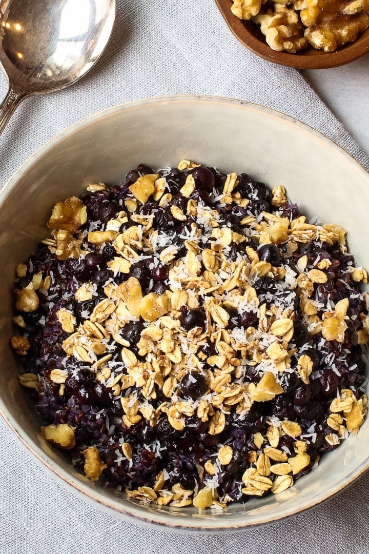 close up of quinoa porridge with black lentils and blueberries, topped with granola and shredded coconut in a bowl on a blue towel with a spoon and a small bowls of walnuts.